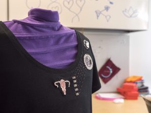 black t-shirt with an embroidered uterus on the chest; 5 pink LEDs are mounted on the shirt next to the uterus. The LEDs are connected to a LilyPad Arduino by conductive thread. The background shows a slightly messy workshop. 