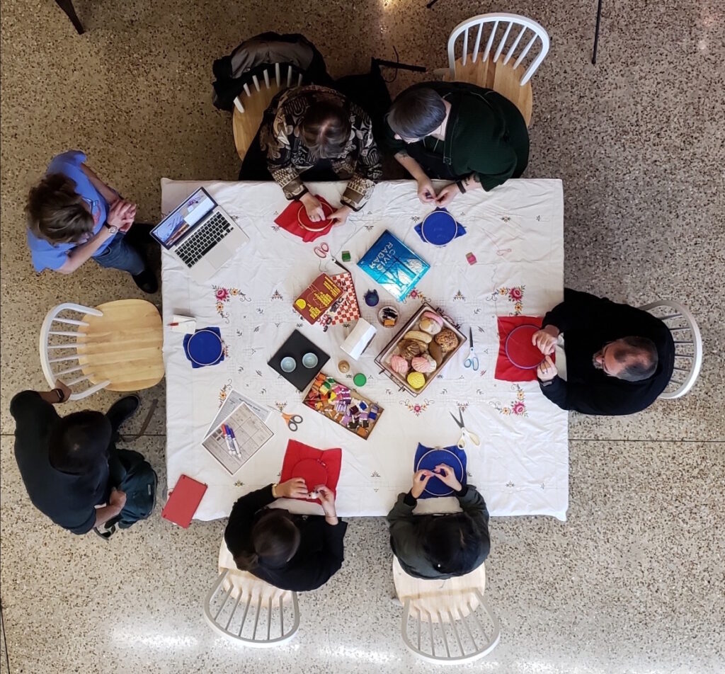 A kitchen table shown from above. People of various ages, races, and genders sit at the table working on embroidery hoops. The table includes books, pan dulce, and other remnants of our childhoods. 